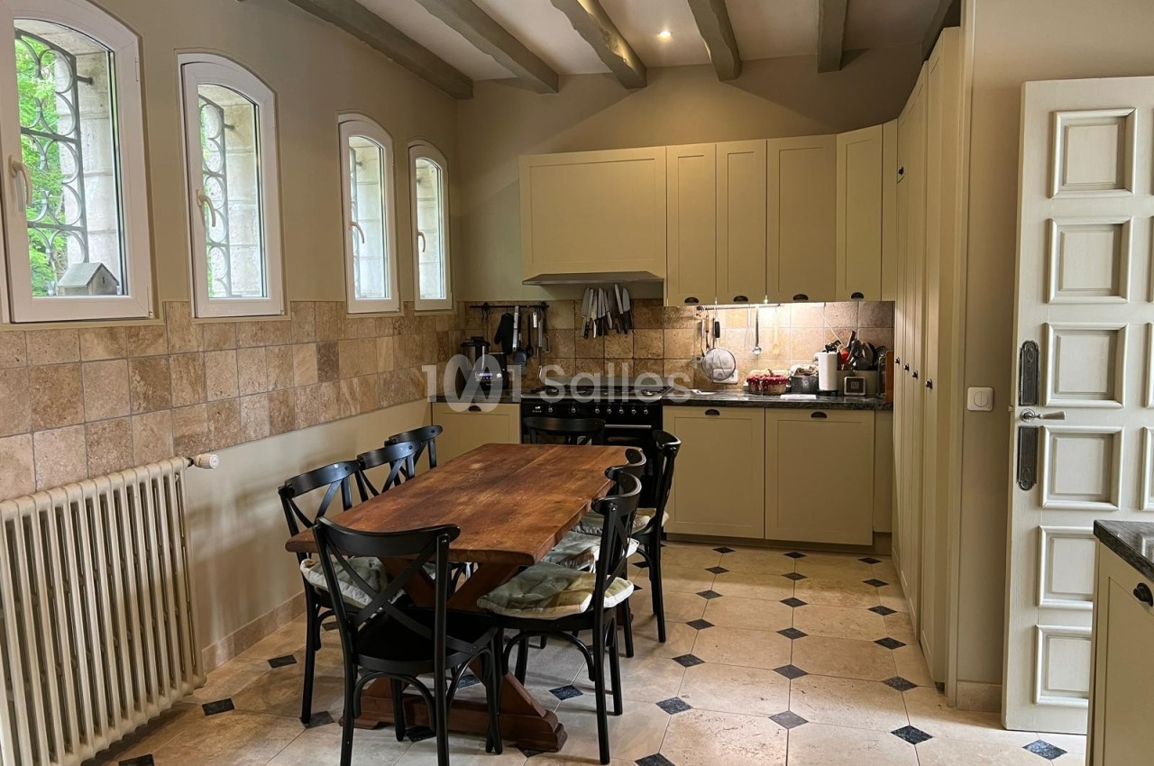Cuisine lumineuse avec table en bois, chaises noires, meubles beige clair, carrelage clair et fenêtres en arc.
