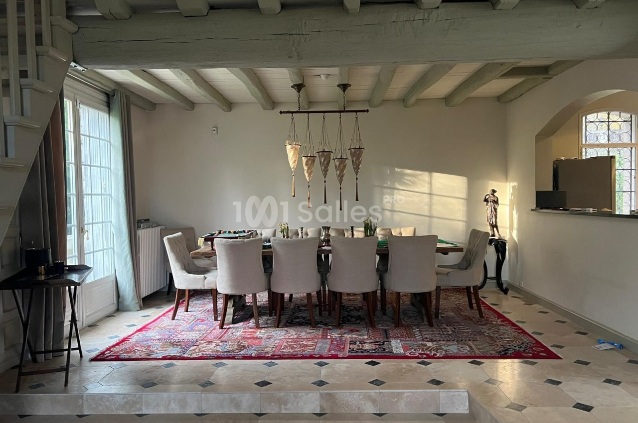 Salle à manger lumineuse avec une grande table entourée de chaises, tapis rouge au sol et poutres apparentes au plafond.