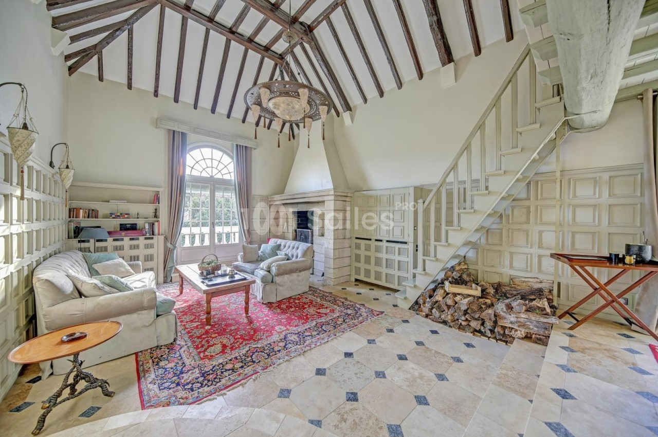Salon lumineux avec poutres apparentes, cheminée en pierre, escalier en bois et mobilier classique sur tapis coloré.
