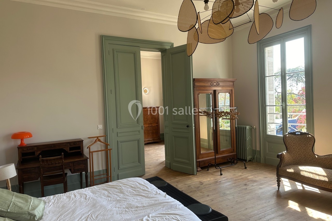 Chambre lumineuse avec lit, bureau en bois, armoire ancienne, chaise et vue sur l'extérieur par une grande fenêtre.