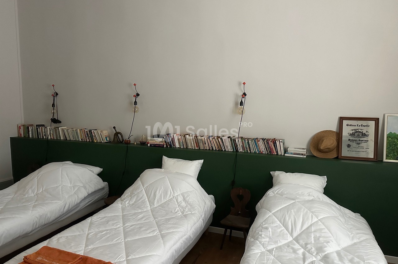 Trois lits simples alignés avec des couvertures blanches, une étagère murale avec des livres et des lampes suspendues.