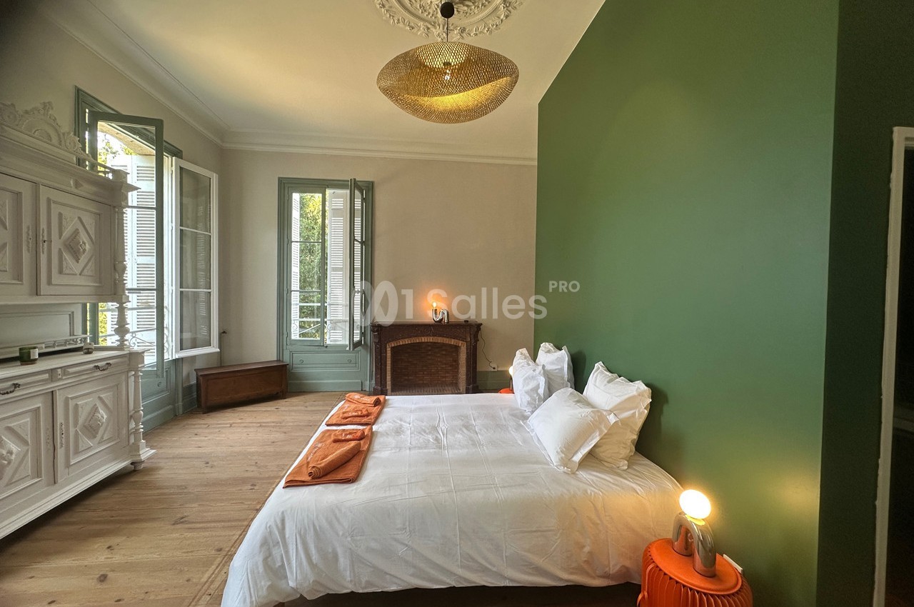 Chambre lumineuse avec murs verts, lit double, cheminée ancienne, mobilier blanc et grandes fenêtres ouvertes.