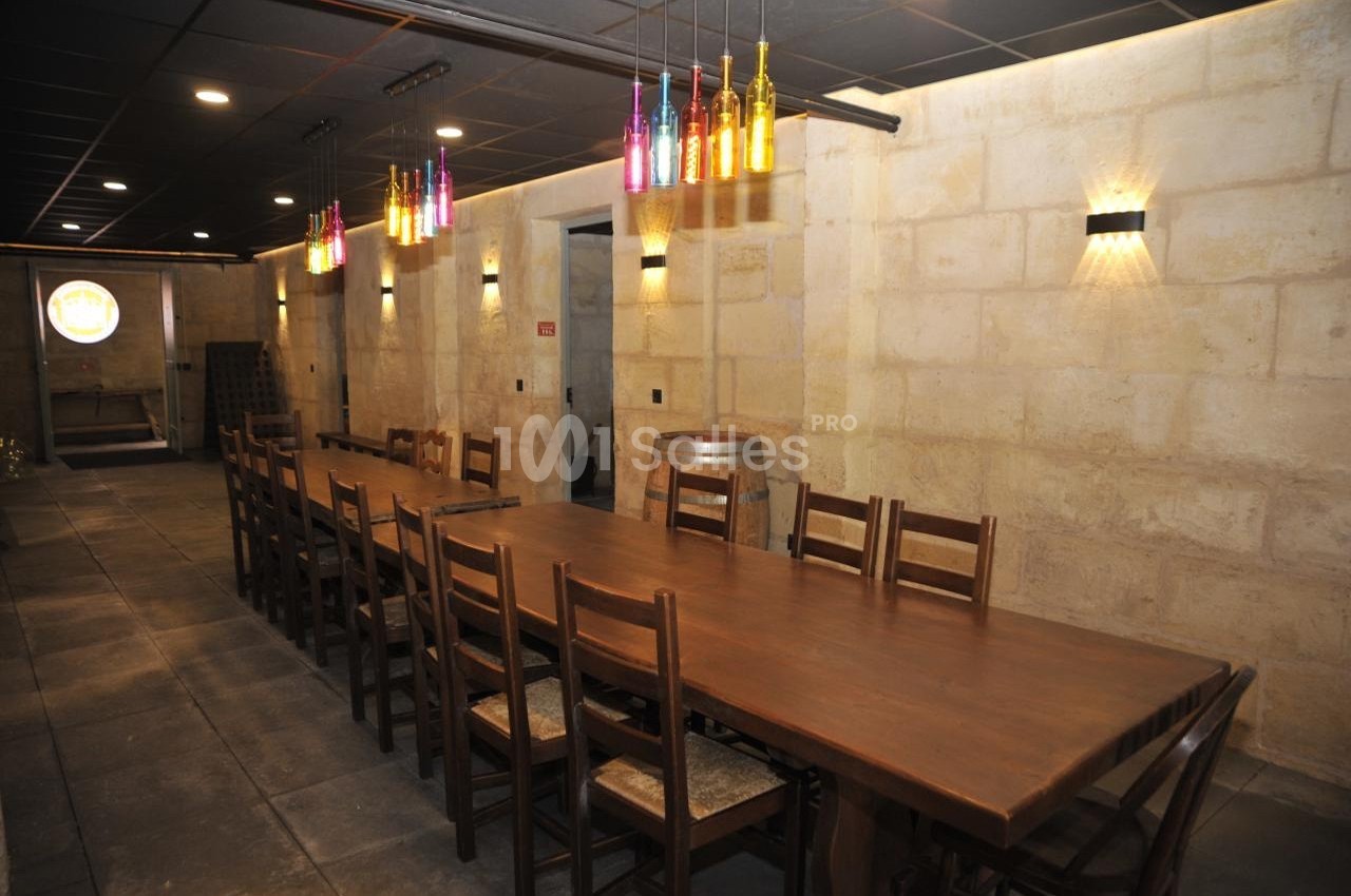 Salle avec une longue table en bois entourée de chaises, éclairée par des lampes murales et des bouteilles suspendues.