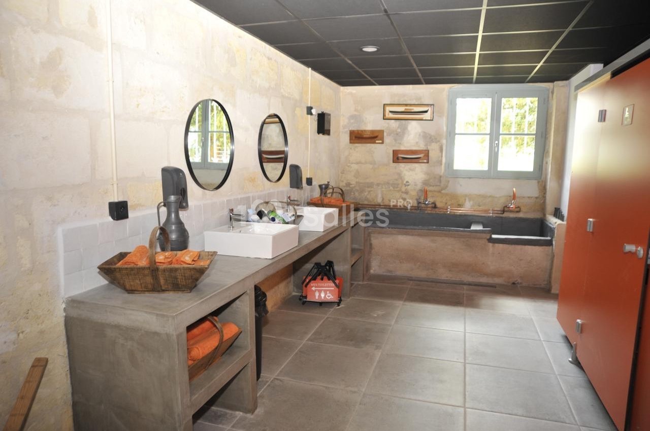 Salle de bain avec murs en pierre, deux lavabos, miroirs ronds, baignoire, et rangements en béton brut.