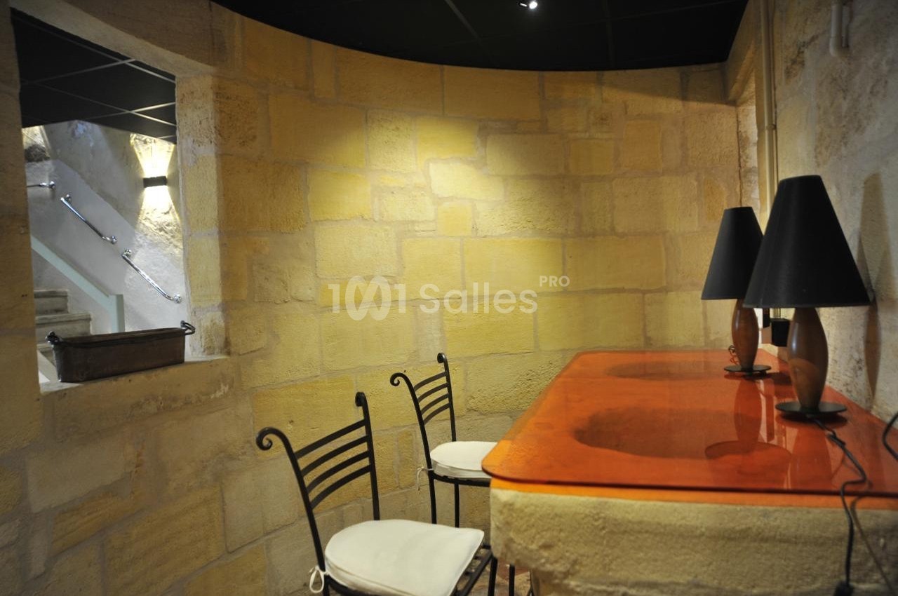 Mur en pierre apparente avec un comptoir en bois verni orange, deux chaises en métal et des lampes noires.