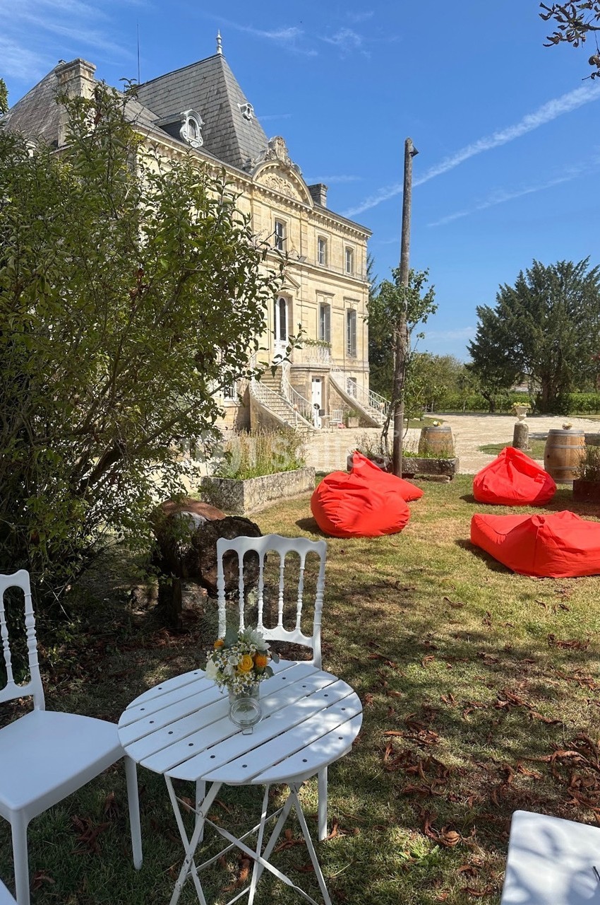 Chaises blanches et table ronde dans un jardin, avec des poufs rouges et un bâtiment ancien en arrière-plan.