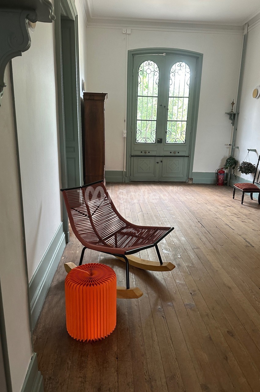 Chaise à bascule en bois et métal avec un tabouret orange dans un couloir lumineux au sol en parquet.