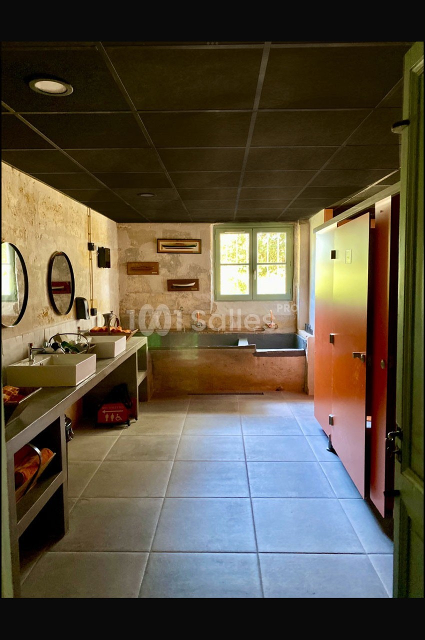 Salle de bain lumineuse avec murs en pierre, lavabos modernes, cabines orange et fenêtre laissant entrer la lumière…