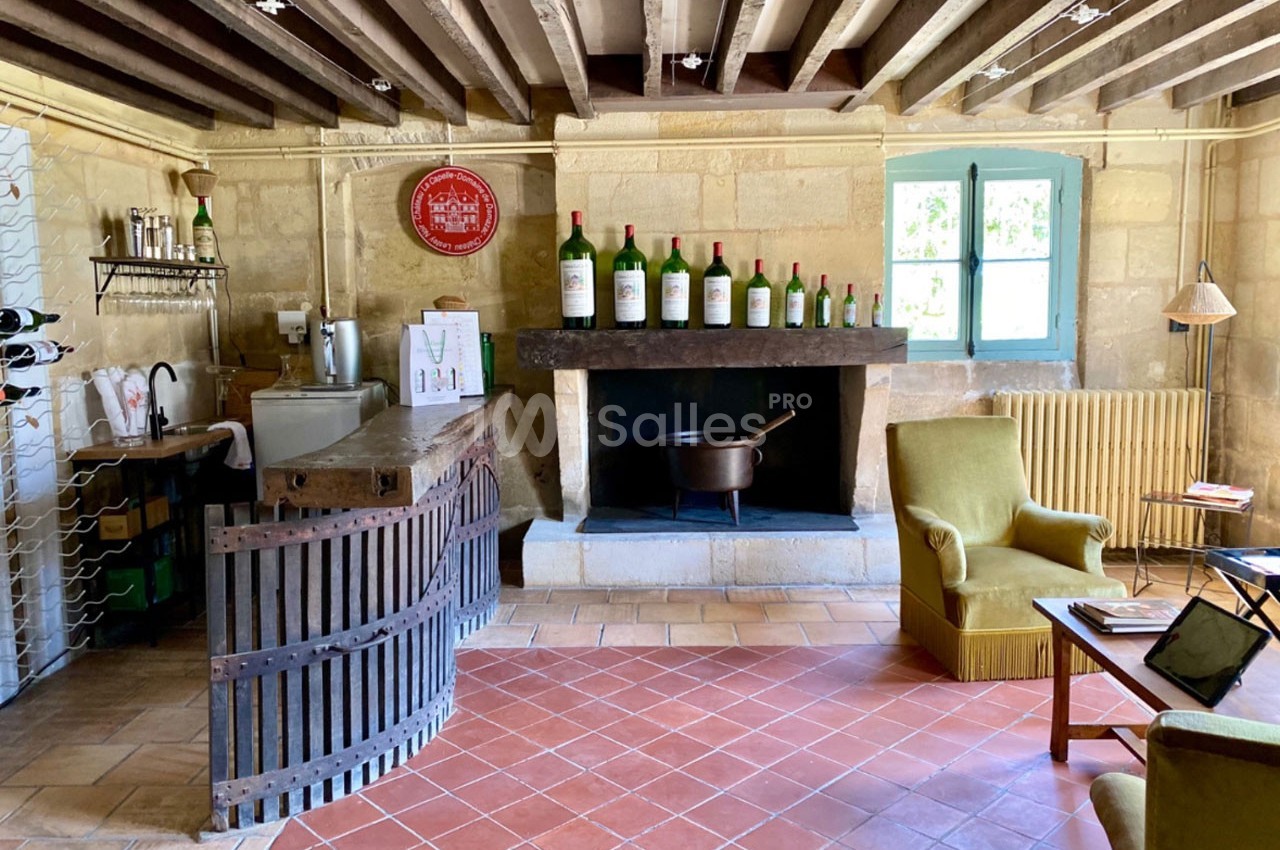Salle rustique avec cheminée, bar en bois, bouteilles de vin alignées et fauteuils près d'une fenêtre lumineuse.
