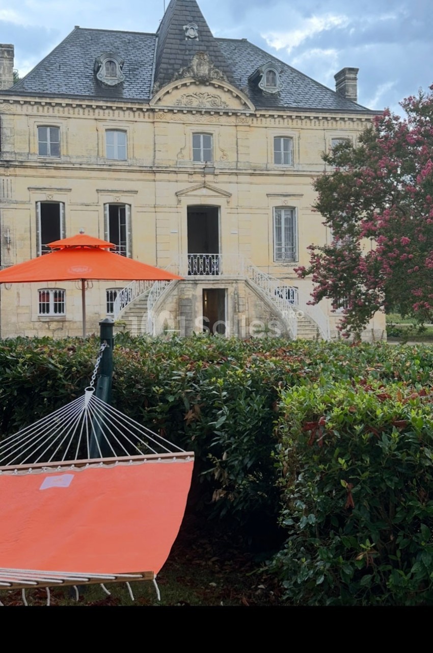 Hamac orange installé dans un jardin devant une grande maison en pierre avec un toit mansardé et des arbres fleuris.