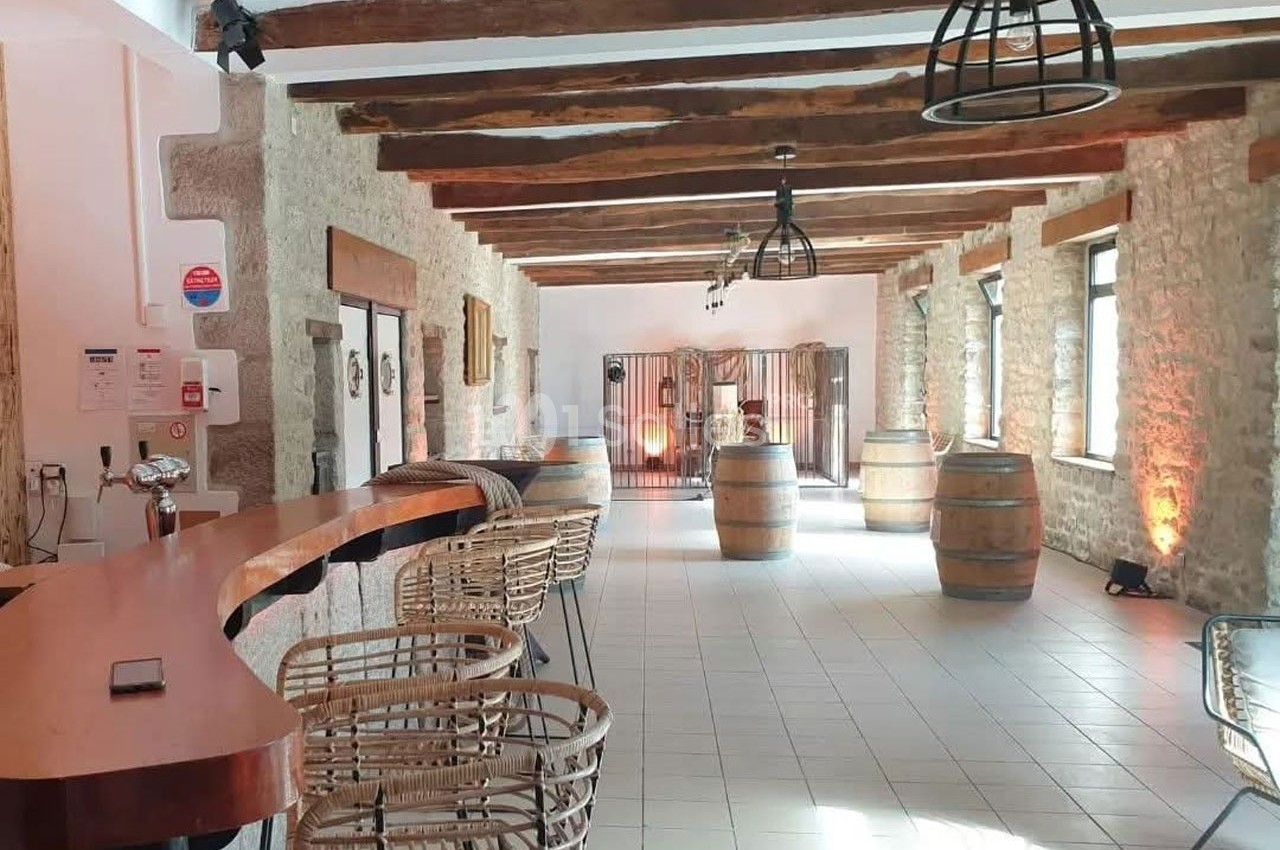 Salle lumineuse avec murs en pierre, poutres apparentes, bar en bois, tonneaux décoratifs et chaises en rotin.