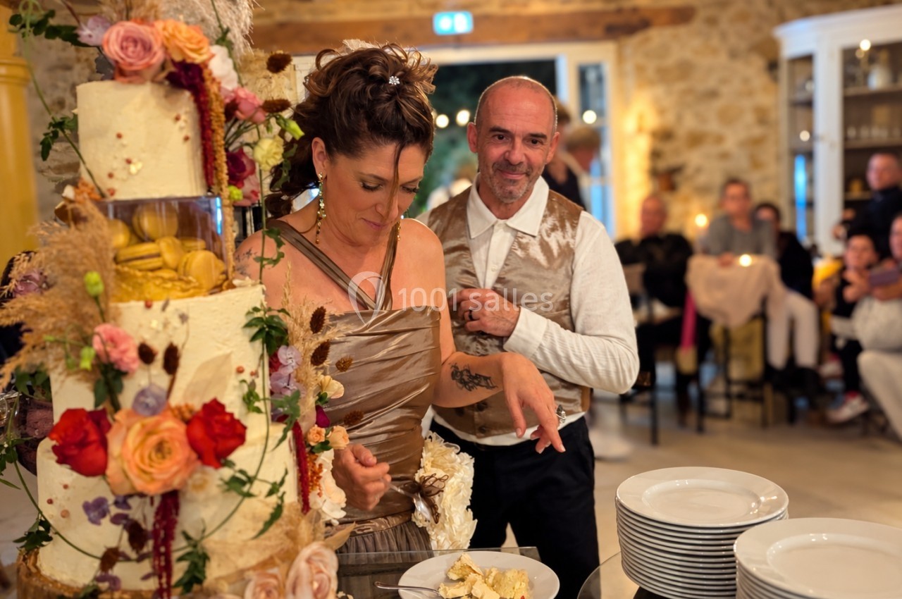 Une femme et un homme près d'un gâteau de mariage décoré, elle sert une part, entourés d'invités dans une salle festive.