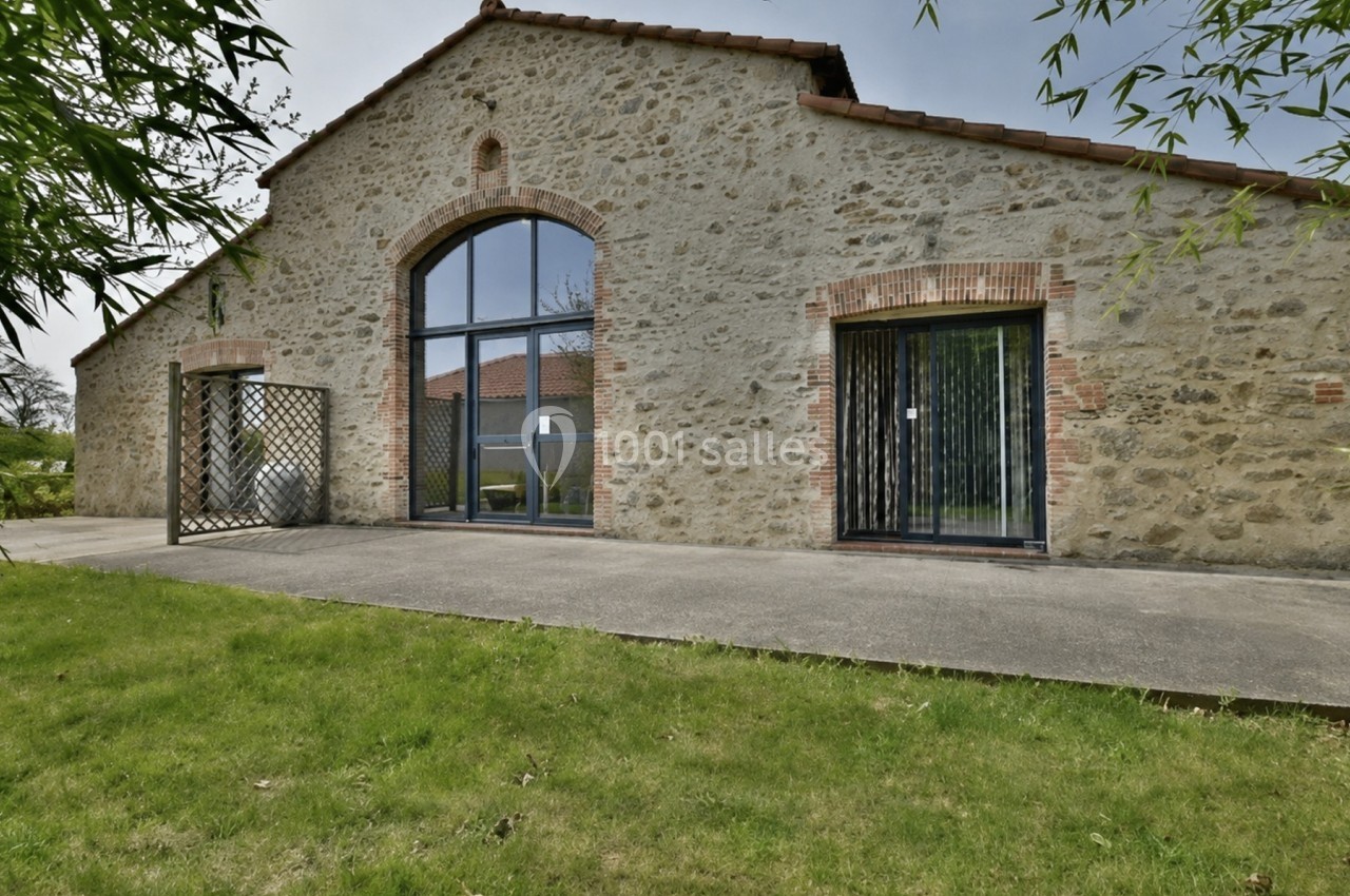 Façade d'une maison en pierre avec de grandes baies vitrées, entourée de pelouse et de végétation.