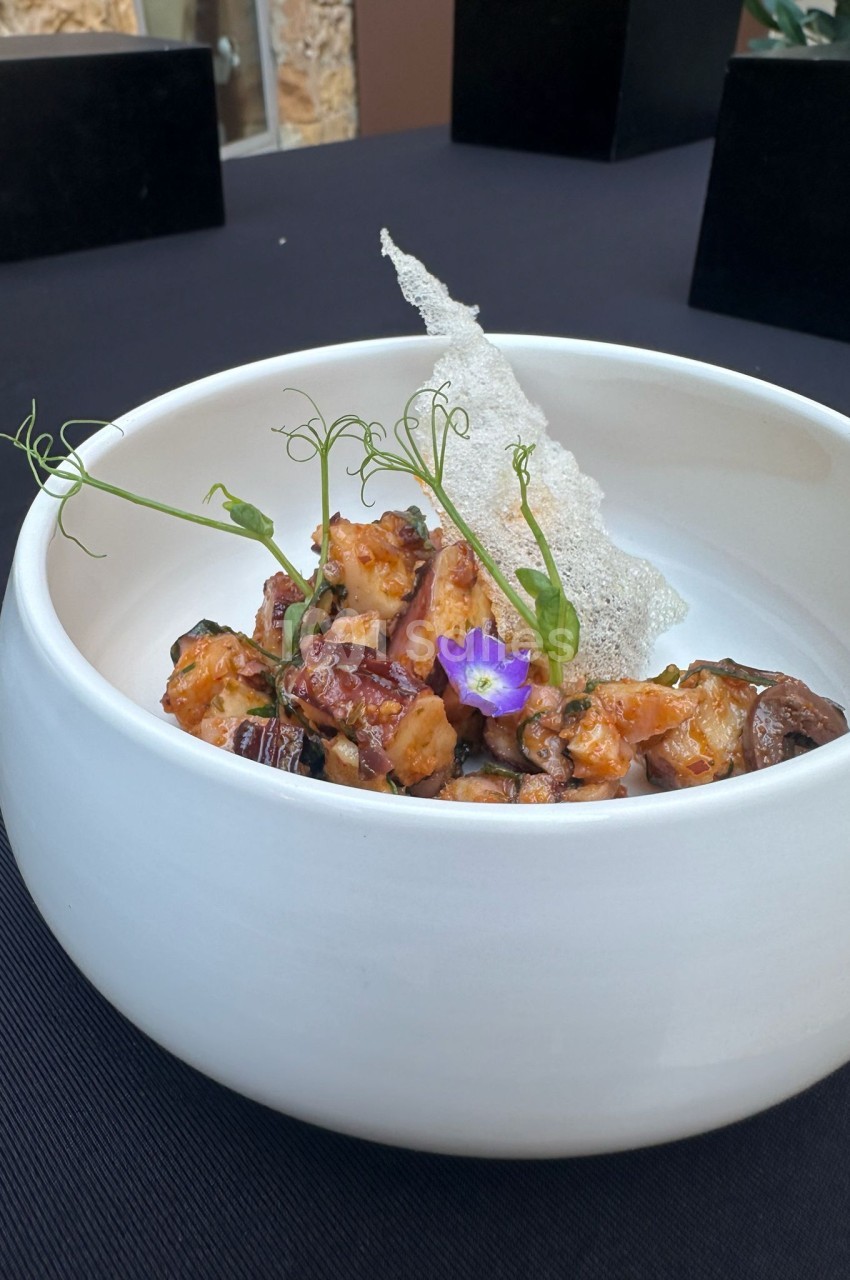 Assiette blanche avec dés de viande braisée, tuile croustillante, micro-herbes et fleur comestible violette.