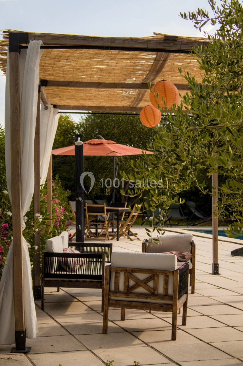 Pergola en bois avec rideaux blancs, mobilier de jardin et lanternes suspendues, près d'une piscine entourée de végétation.