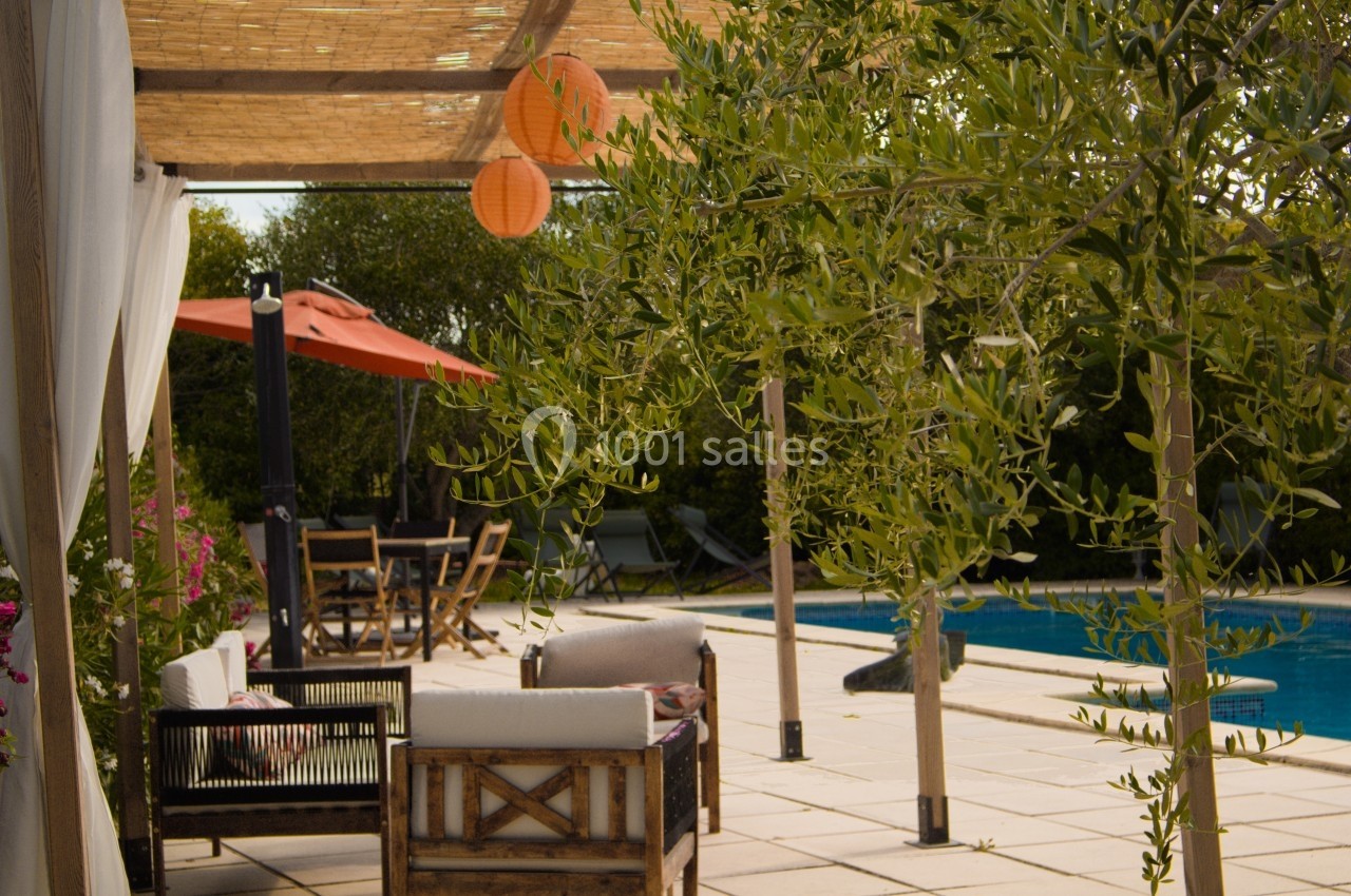 Terrasse ombragée avec salon de jardin en bois, piscine, oliviers et lampions orange sous une pergola.