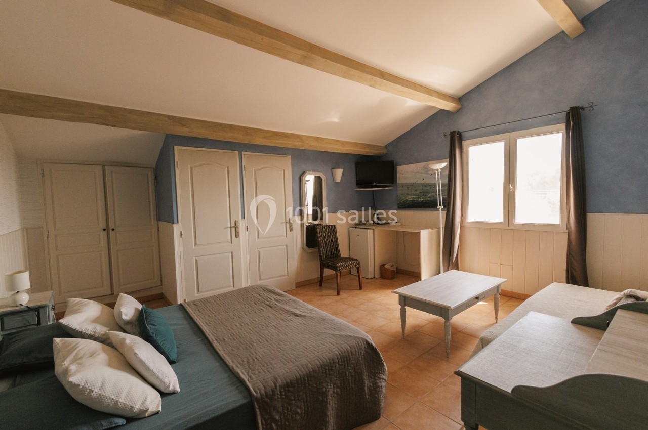 Chambre lumineuse avec poutres apparentes, lit double, lit simple, armoire, bureau et fenêtres donnant sur l'extérieur.