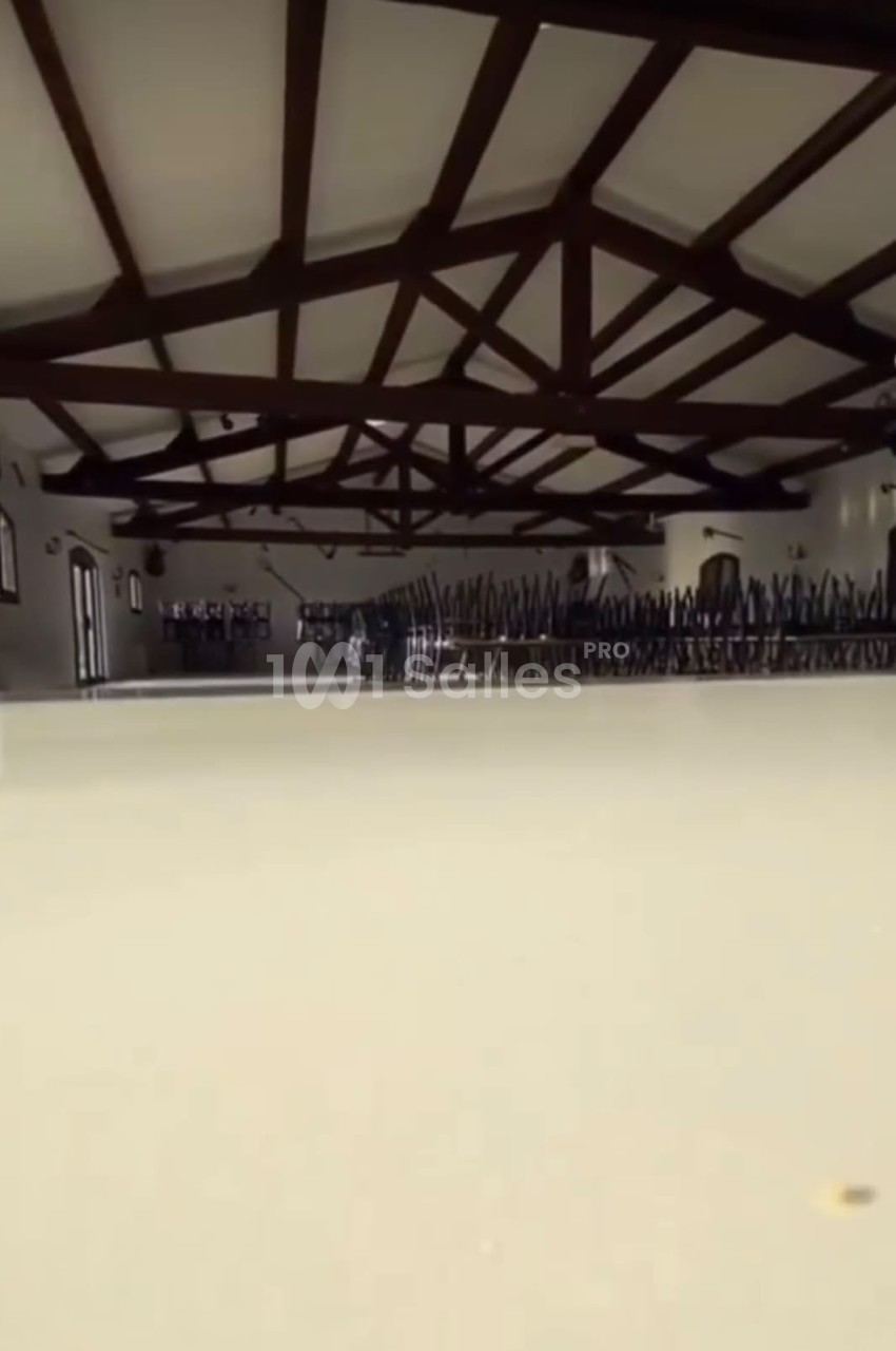 Salle vide avec plafond en poutres apparentes, chaises empilées dans le fond et lumière naturelle entrant par les fenêtres.