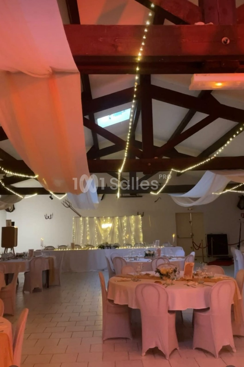 Salle de réception décorée avec des guirlandes lumineuses, des nappes blanches et des tables dressées pour un événement.