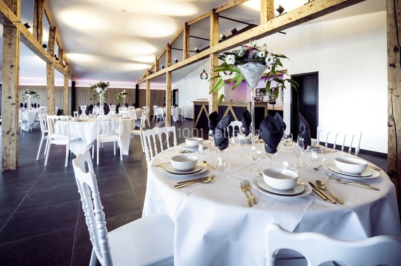 Salle de réception élégante avec tables rondes dressées, nappes blanches, serviettes noires et décorations florales.