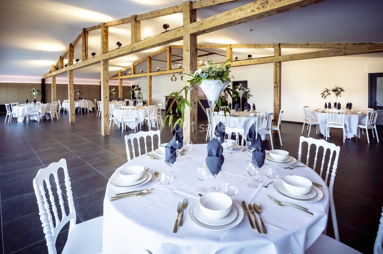 Salle de réception lumineuse avec tables rondes dressées, décorée de plantes et poutres apparentes en bois.