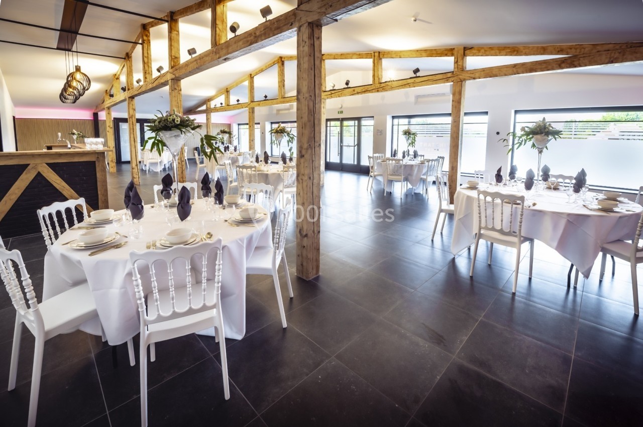 Salle de réception lumineuse avec tables rondes dressées, chaises blanches et poutres apparentes en bois.