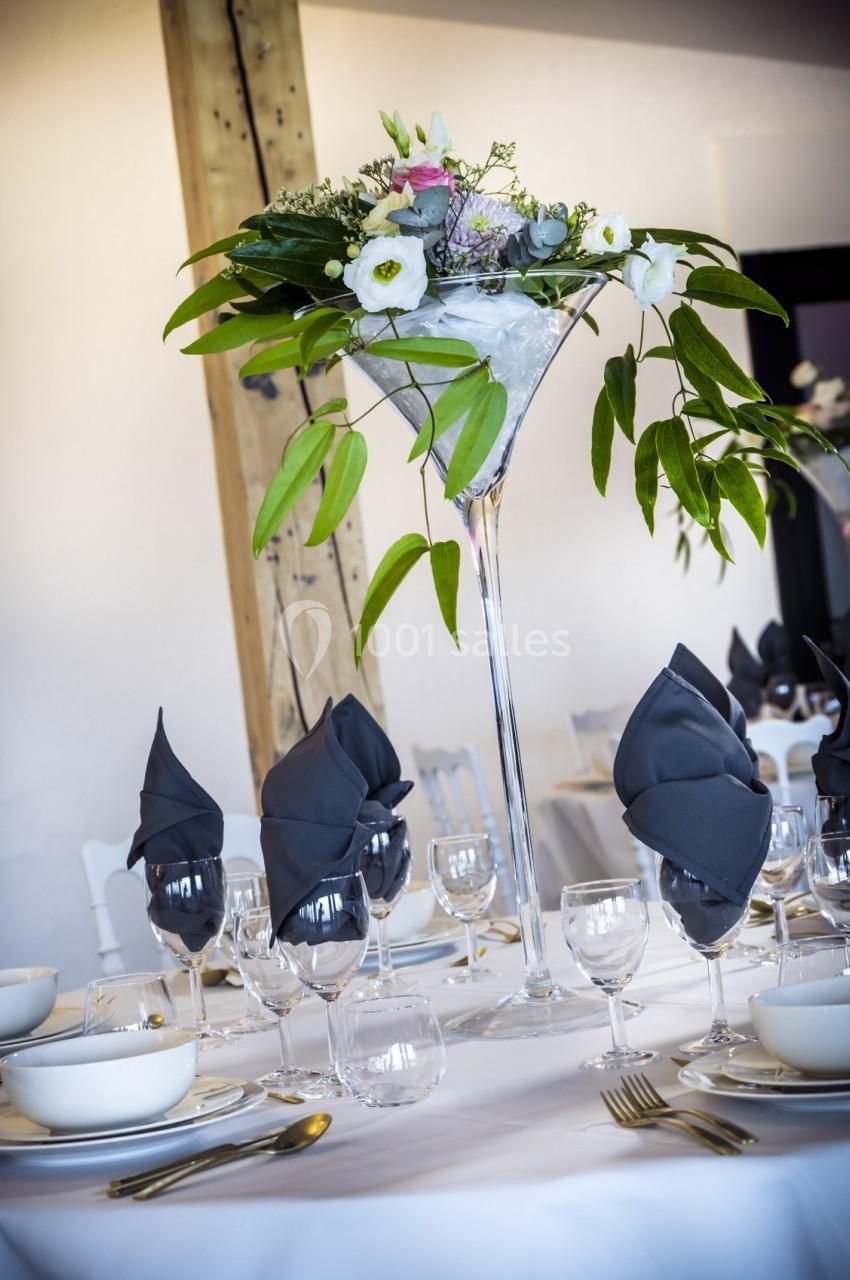 Centre de table floral dans un vase haut, entouré de vaisselle élégante et serviettes pliées sur une table dressée.