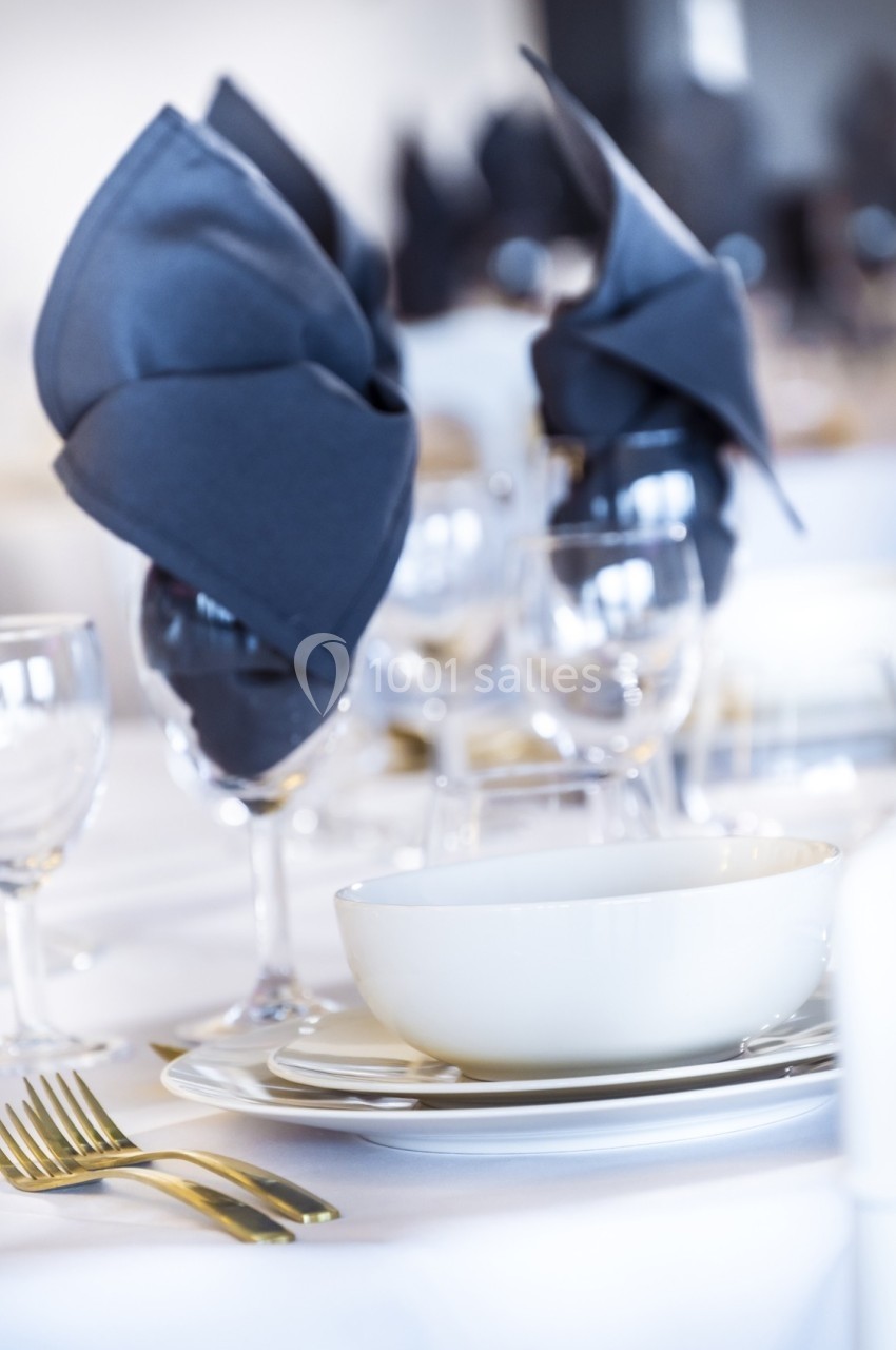 Table dressée avec assiette blanche, couverts dorés, verres et serviettes pliées en éventail bleu foncé.
