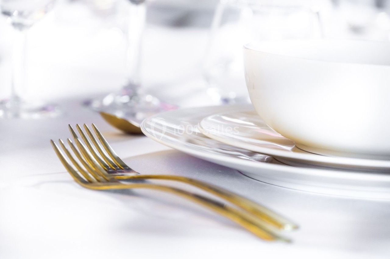 Assiette blanche avec bol assorti, couverts dorés et verres en arrière-plan sur une table dressée.