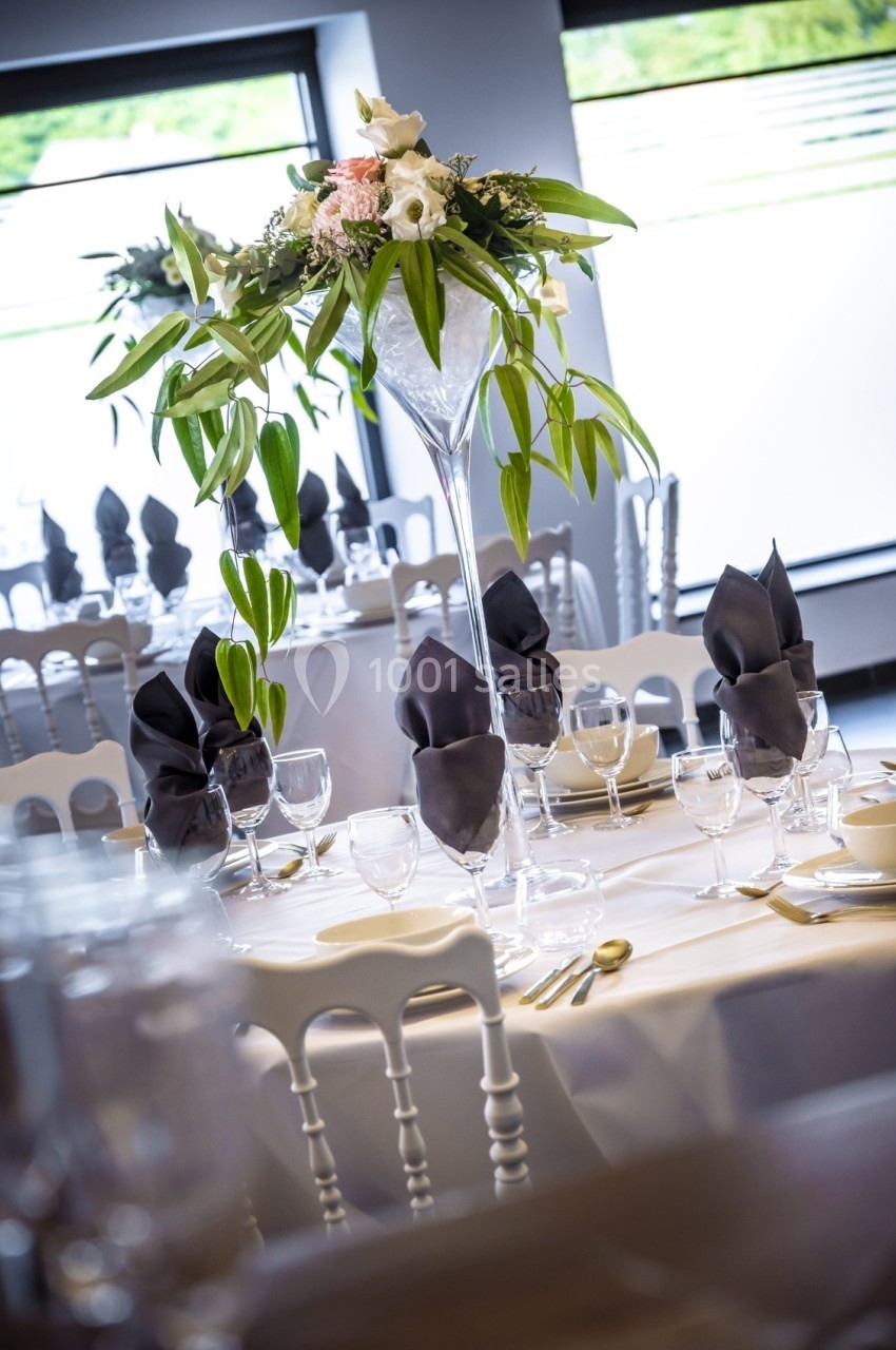 Table élégamment dressée avec nappes blanches, serviettes noires pliées et centre de table floral dans un vase haut.