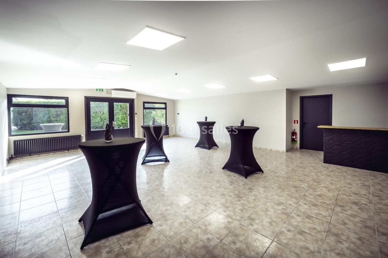 Salle lumineuse avec tables hautes noires, sol carrelé, grandes fenêtres et porte donnant sur un extérieur verdoyant.