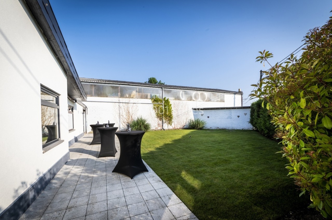 Jardin avec pelouse, terrasse en dalles claires, haies et murs blancs sous un ciel dégagé.