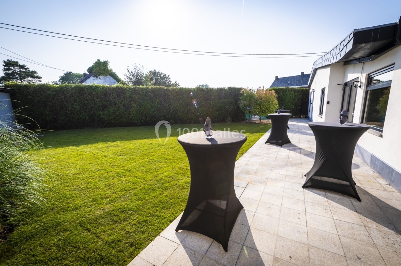 Tables hautes noires sur une terrasse pavée, bordée d'une pelouse et d'une haie, par une journée ensoleillée.