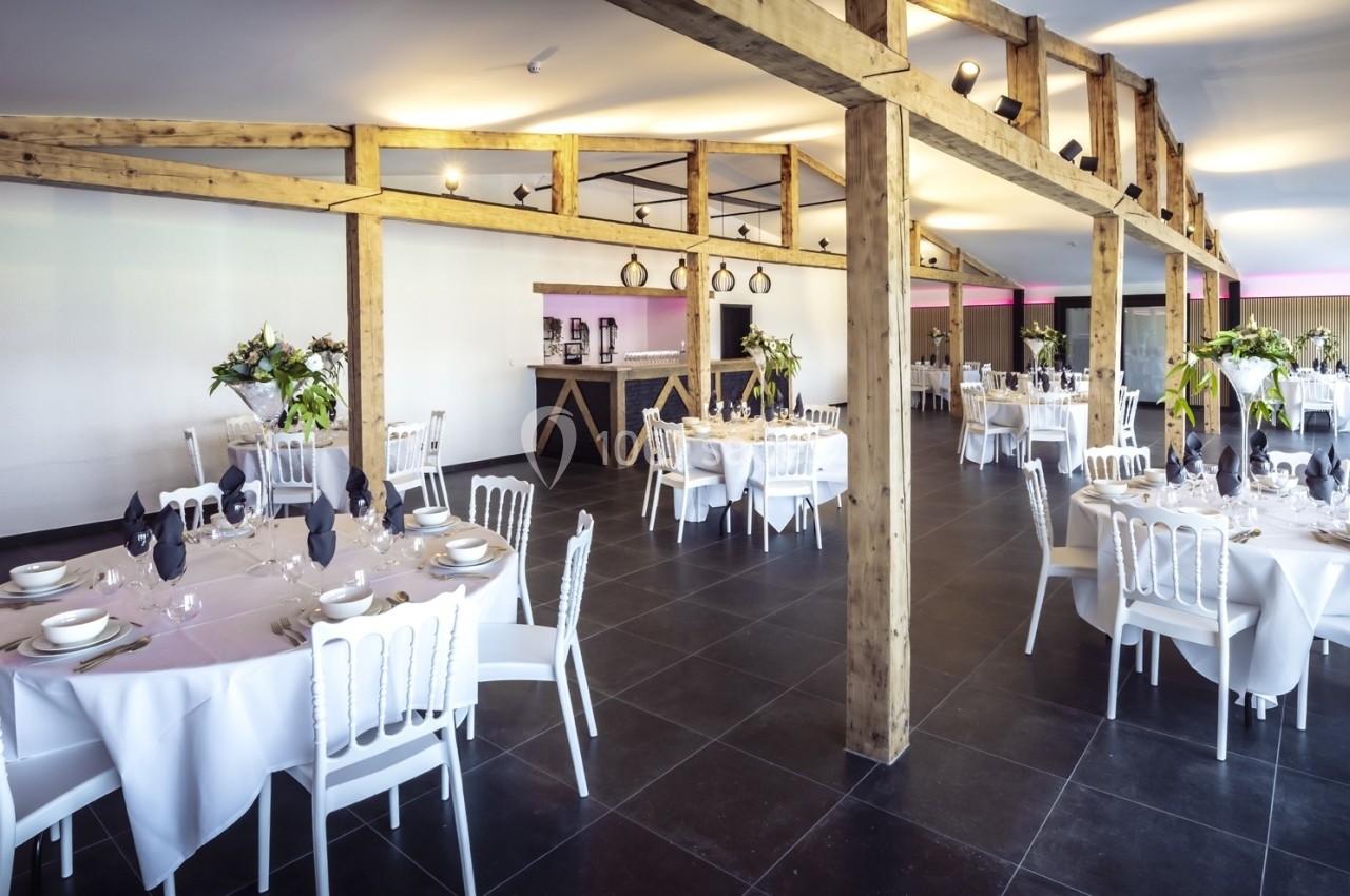 Salle de réception lumineuse avec tables rondes dressées, chaises blanches et poutres en bois apparentes.