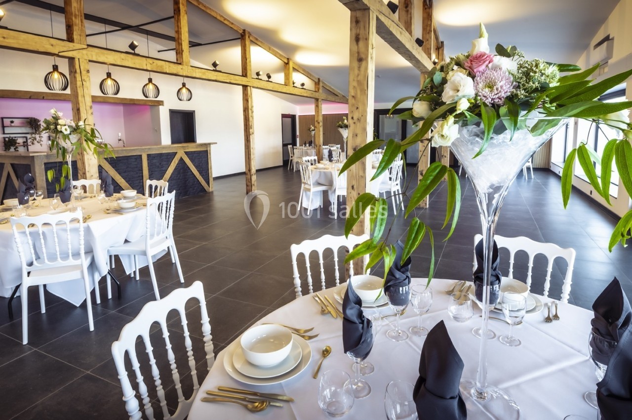 Salle de réception lumineuse avec tables élégamment dressées, décor floral et poutres apparentes en bois.