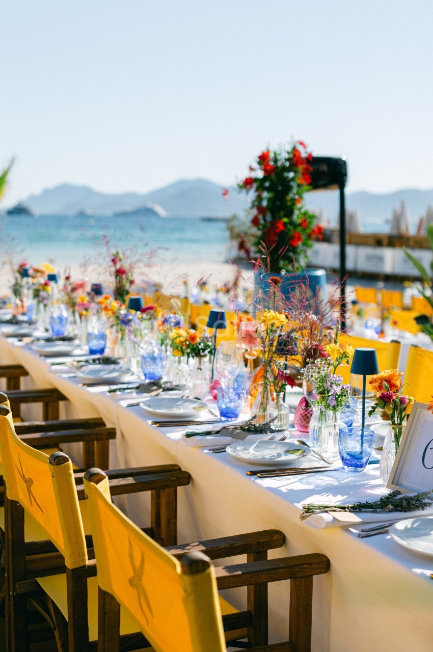 Table dressée en extérieur avec nappes blanches, chaises jaunes et décorations florales colorées, vue sur la plage et la mer.