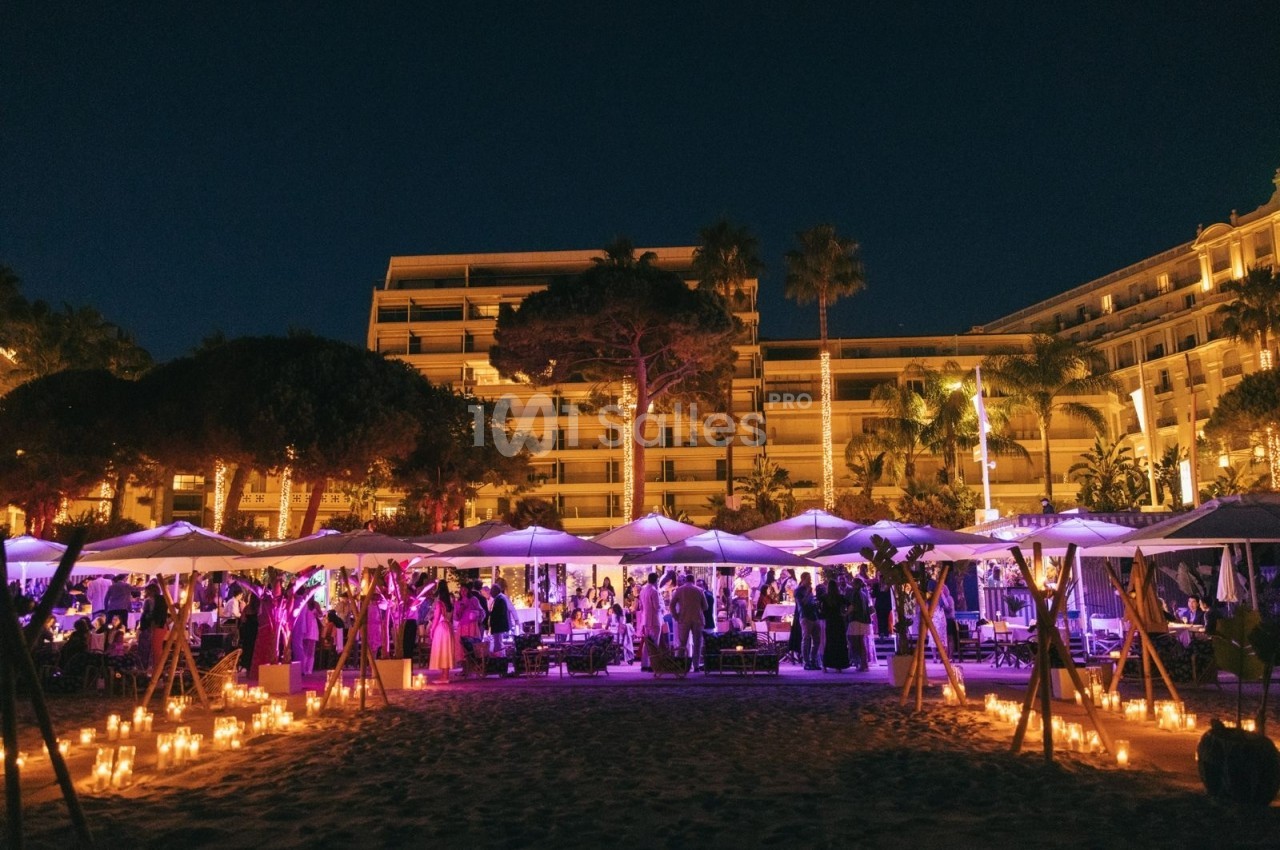 Soirée en plein air sur une plage éclairée par des bougies, avec parasols et un bâtiment illuminé en arrière-plan.