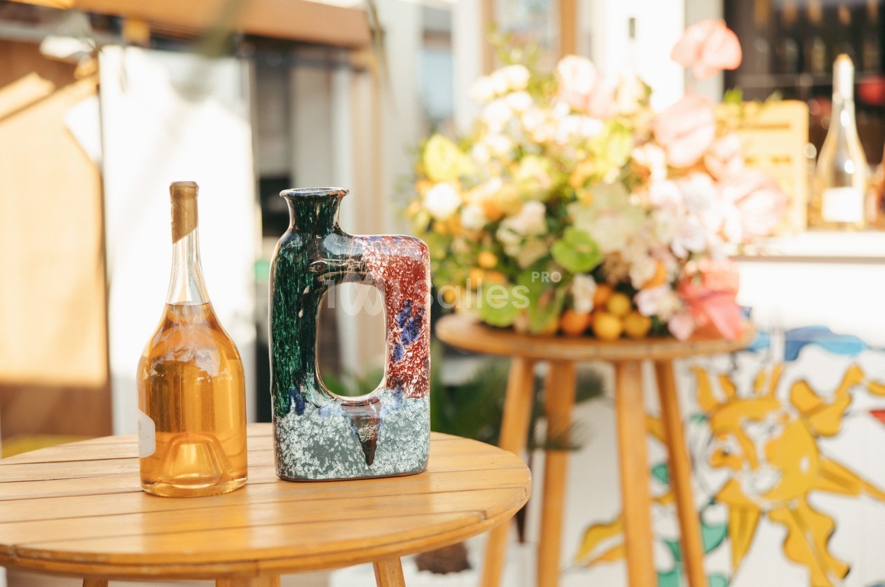 Bouteille de vin et vase coloré posés sur une table en bois, avec un bouquet de fleurs en arrière-plan.