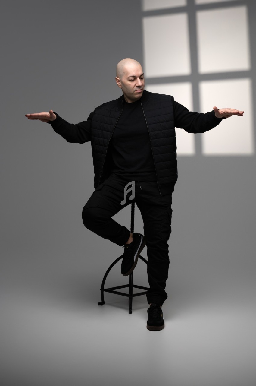 Homme assis sur un tabouret, vêtu de noir, les bras tendus, éclairé par une lumière traversant une fenêtre.