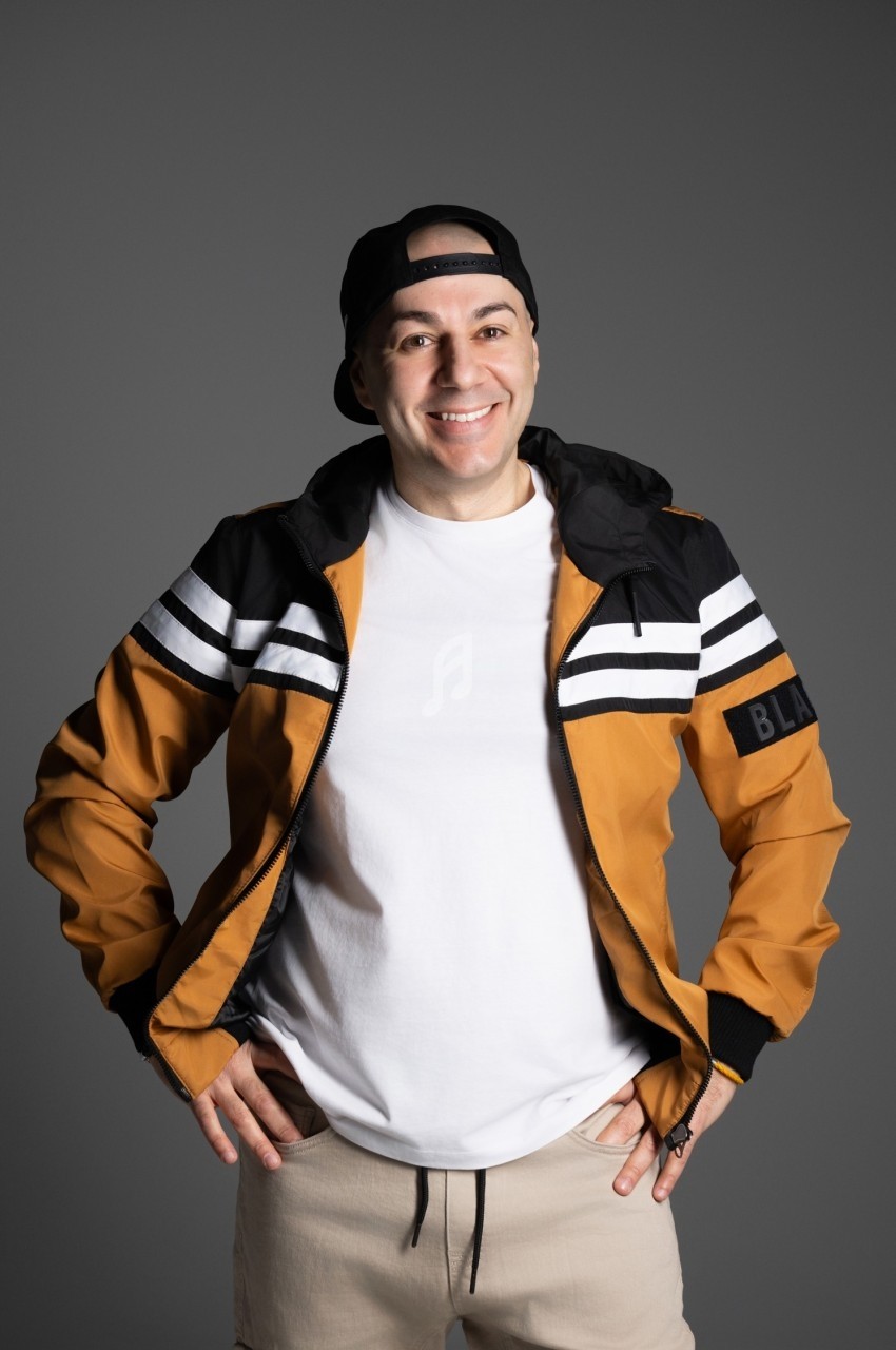 Homme souriant portant une casquette noire, une veste orange à rayures blanches et un t-shirt blanc, sur fond gris.