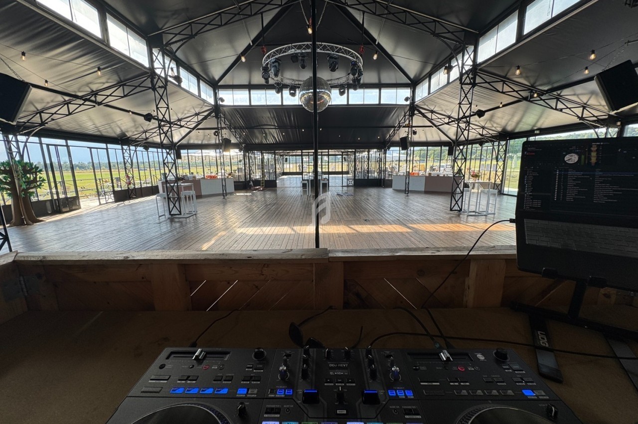Vue depuis une table de mixage sur une grande salle de réception lumineuse avec structure métallique et piste en bois.