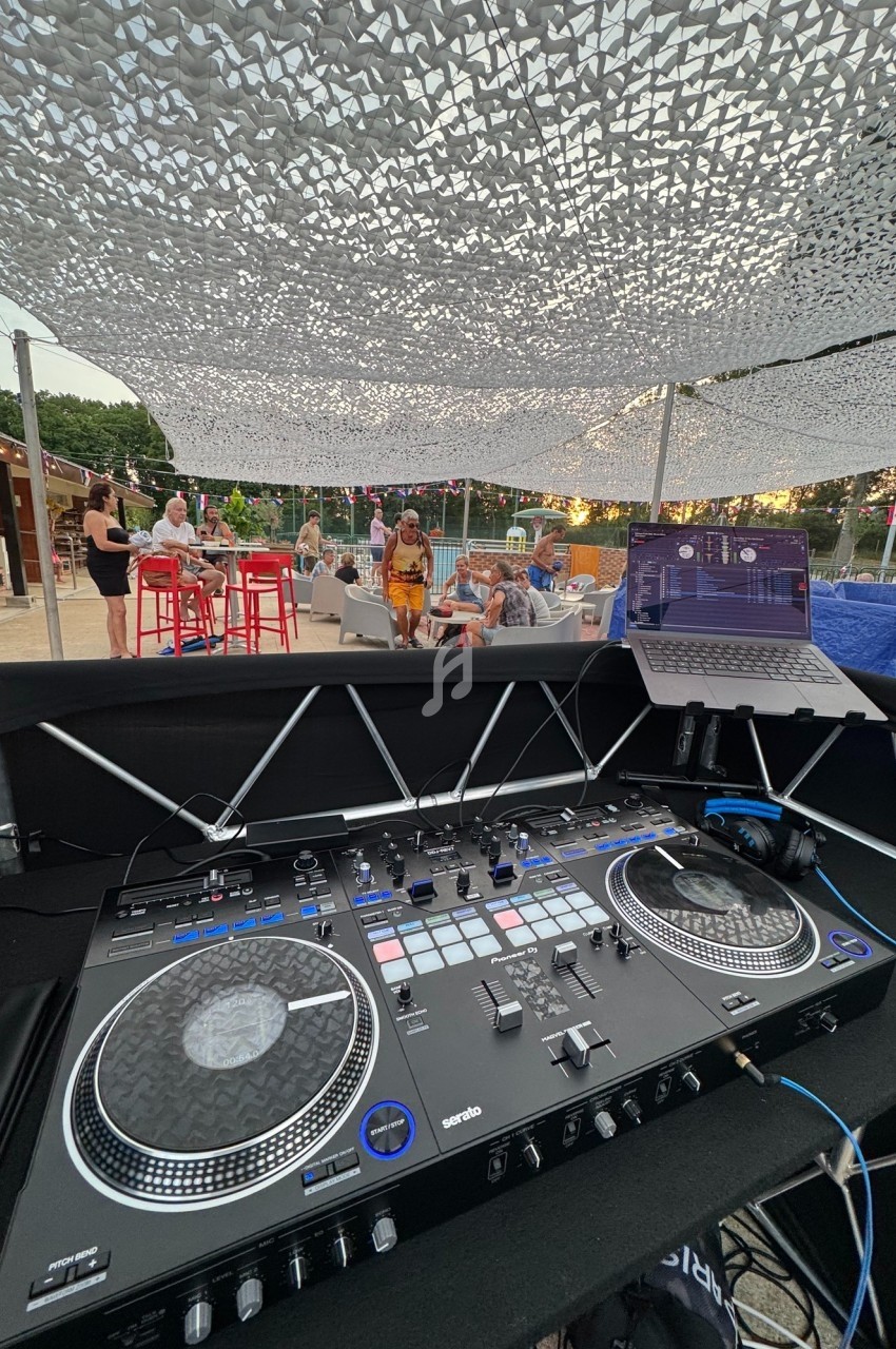 Vue depuis une table de mixage DJ avec ordinateur, sous un filet d'ombrage, en arrière-plan une terrasse animée.