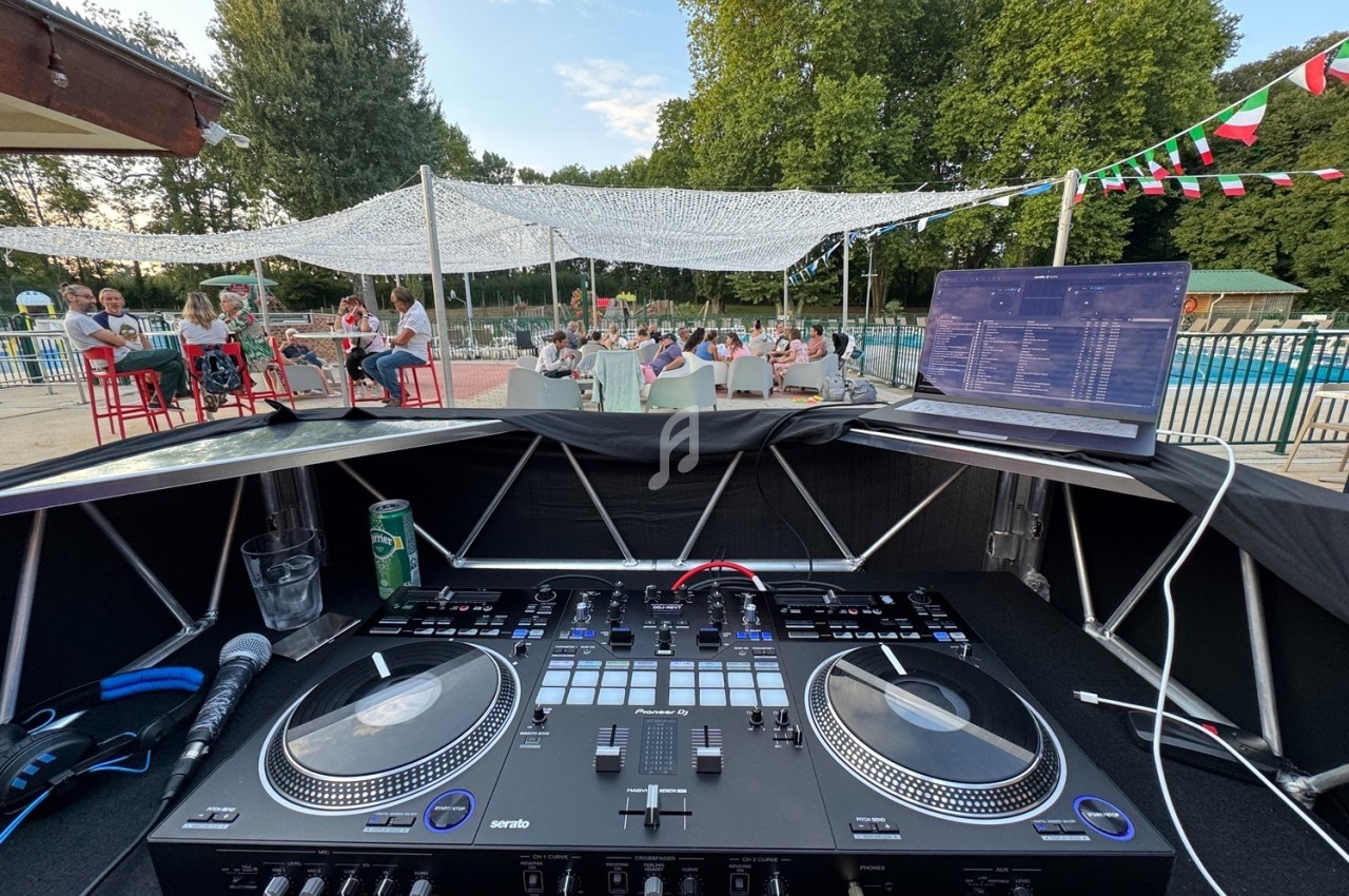 Vue depuis une table de mixage avec ordinateur, micro et boissons, face à une terrasse extérieure animée près d'une piscine.