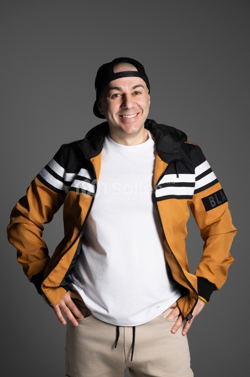 Homme souriant portant une casquette noire, une veste orange à rayures blanches et un t-shirt blanc, sur fond gris.