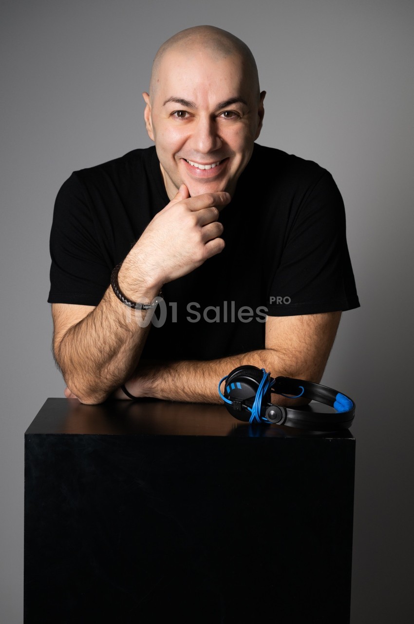 Homme souriant en t-shirt noir, appuyé sur un cube noir avec un casque audio bleu posé devant lui.