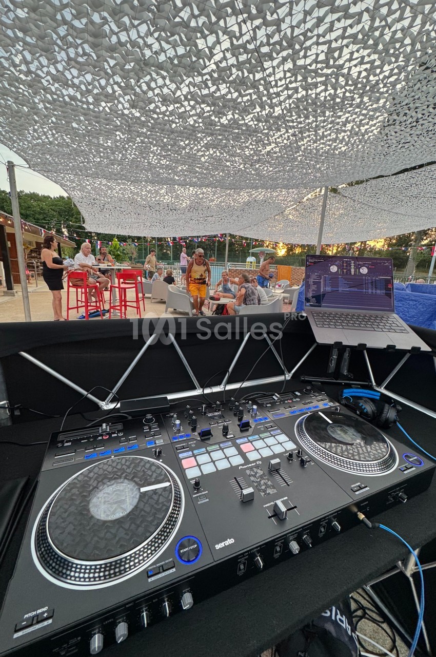 Vue depuis une table de mixage DJ sous un filet d'ombrage, avec un bar et des personnes en arrière-plan.