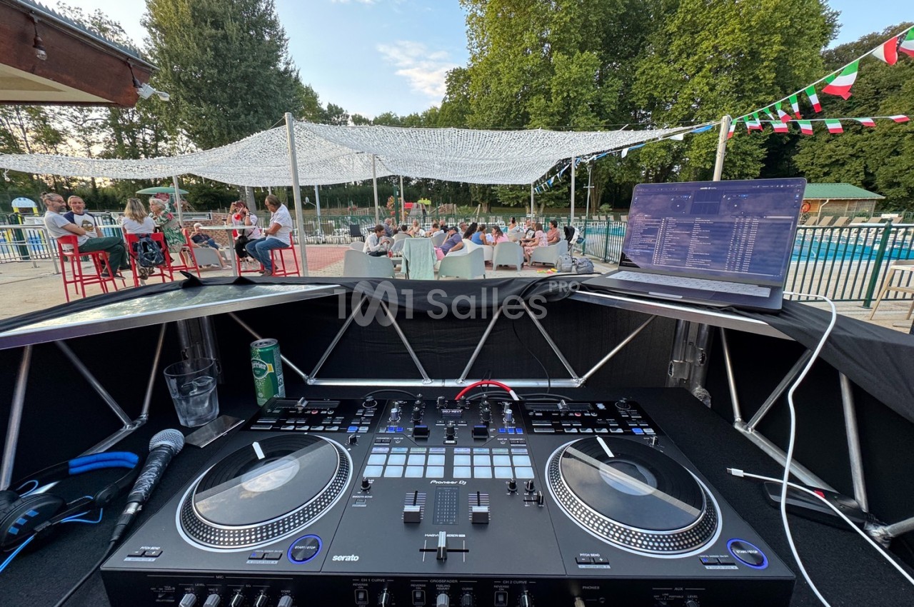 Vue depuis une table de mixage avec ordinateur, micro et boissons, en extérieur près d'une piscine et d'un public assis.