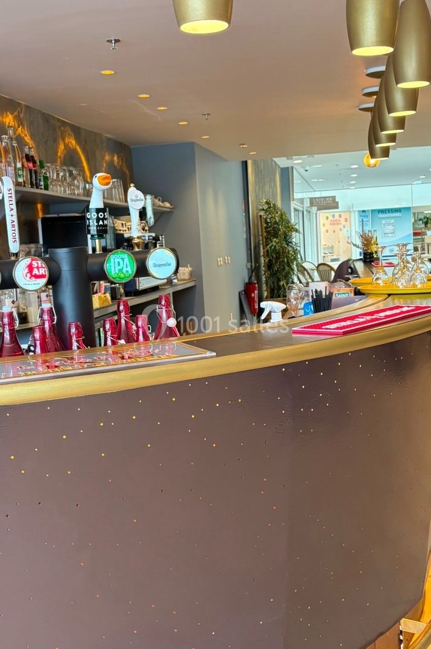 Comptoir de bar moderne avec tireuses à bière, bouteilles en arrière-plan et salle lumineuse en second plan.