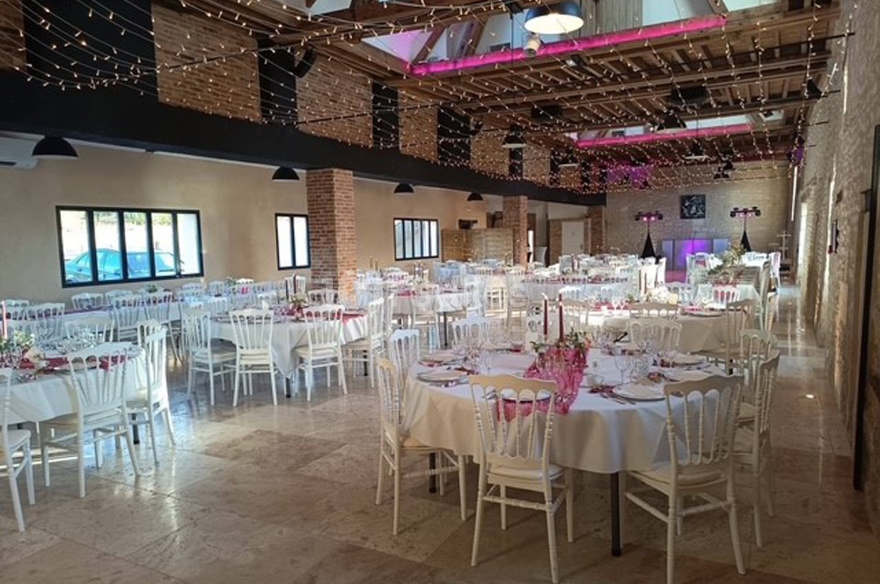 Salle de réception décorée avec des tables rondes dressées, des chaises blanches et des guirlandes lumineuses suspendues.
