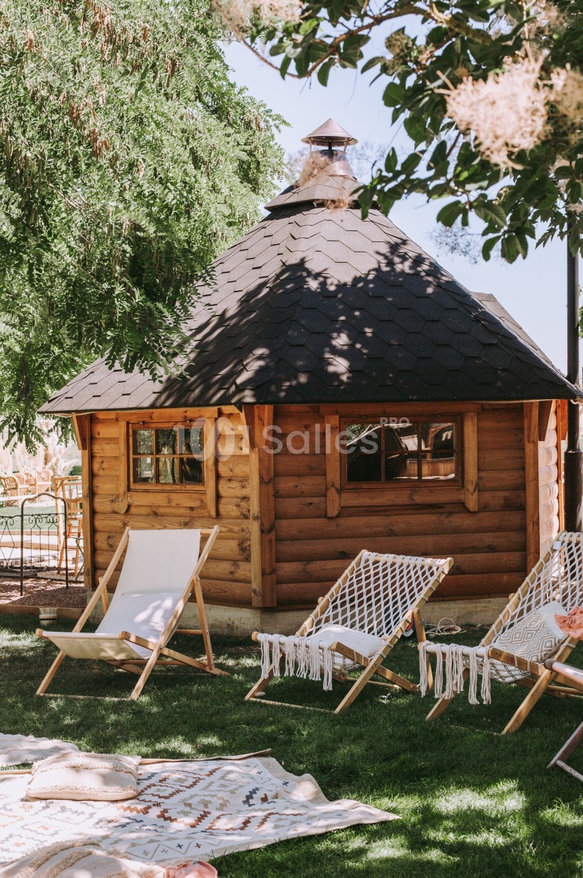 Cabane en bois avec toit en bardeaux entourée de chaises longues et d'arbres dans un jardin ensoleillé.